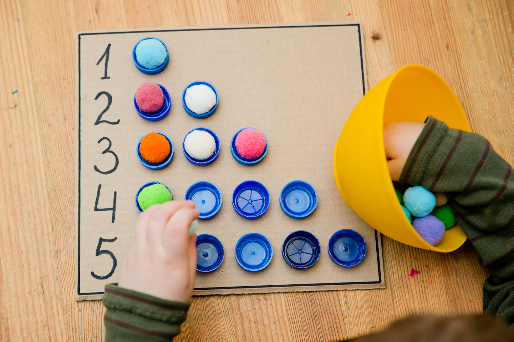  A child is sorting colorful pom poms into bottle caps numbered one to five to learn about numbers.